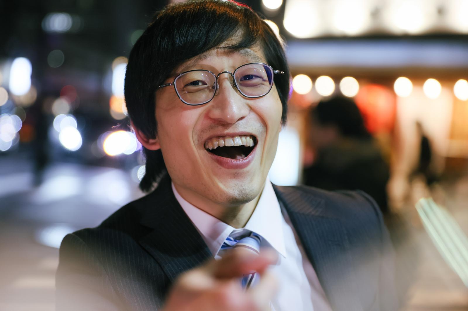 眼鏡をかけた会社員の男性が、繁華街でスーツ姿ではしゃいで笑っている［モデル：あづま］