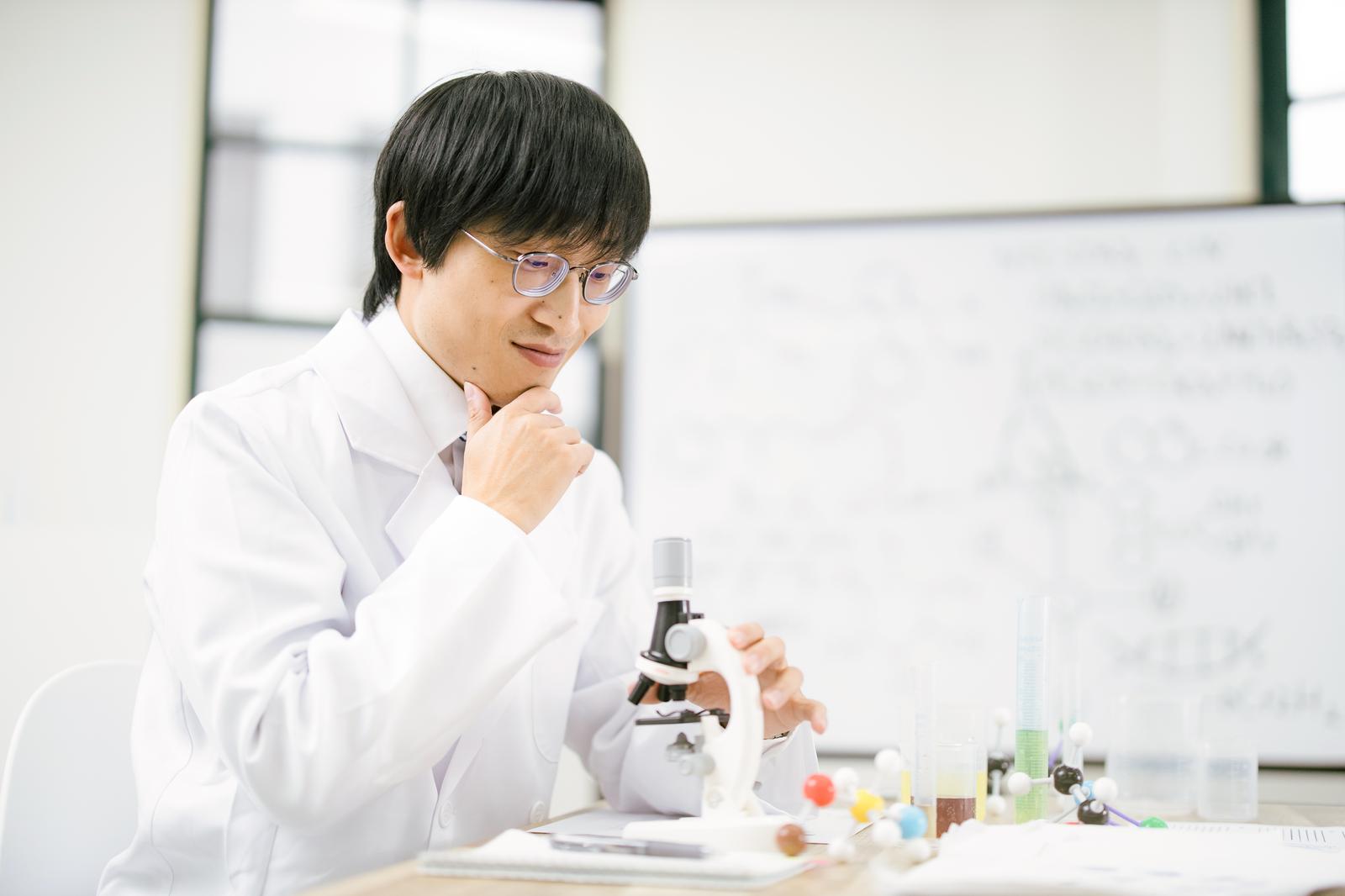 白衣姿で顕微鏡を操作しながら考える男性科学者［モデル：あづま］