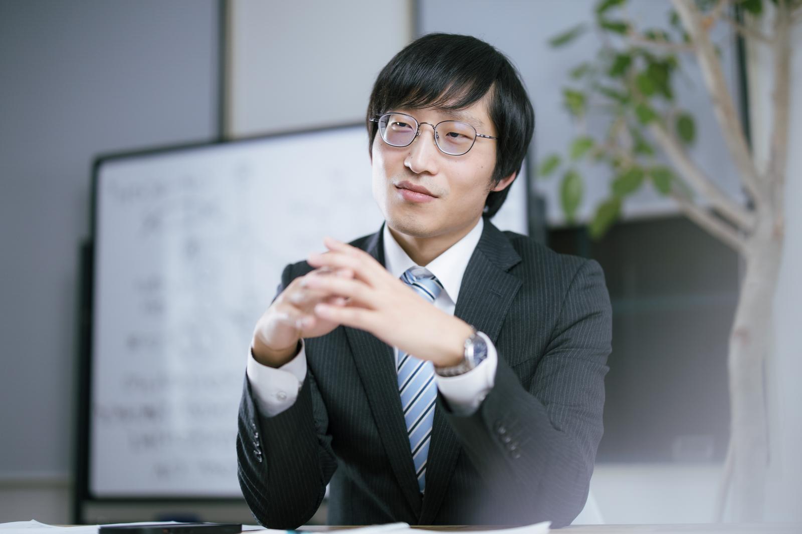 会議室で両手を合わせながら相手の話を聞く眼鏡の男性［モデル：あづま］