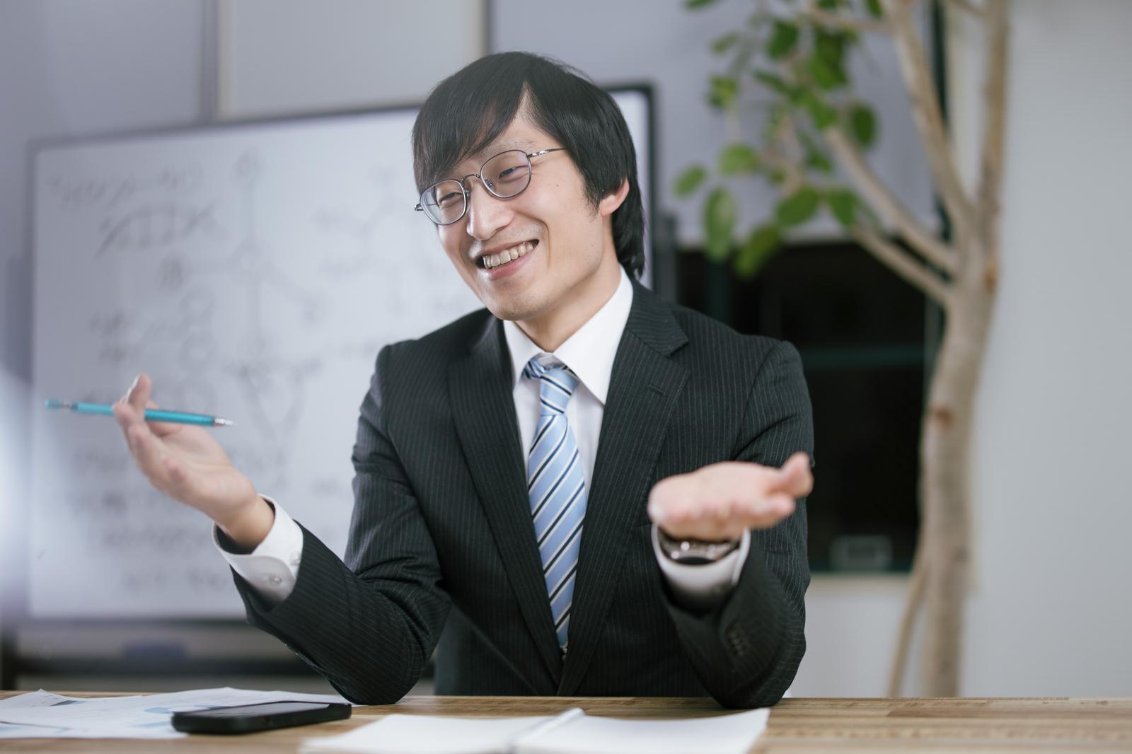 オフィスで机に向かい両手を広げて説明する眼鏡の男性会社員［モデル：あづま］