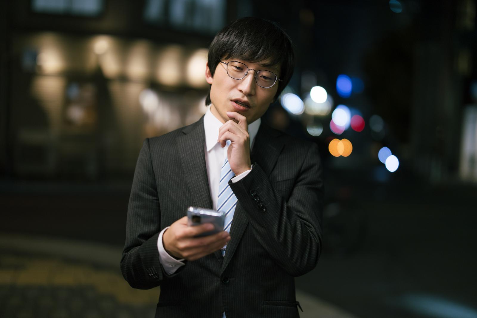 スマートフォンの画面を見て考える眼鏡をかけた黒いビジネススーツの男性［モデル：あづま］