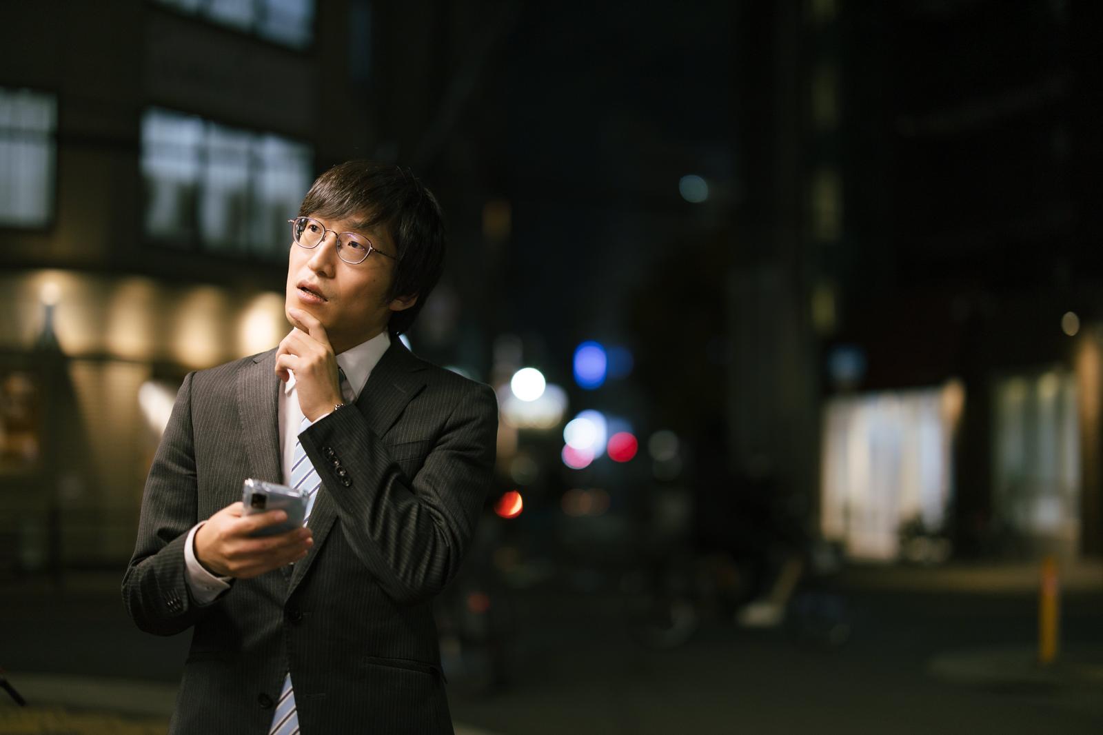 スマートフォンで目的地を検索しながら考える眼鏡をかけたスーツ姿の男性［モデル：あづま］