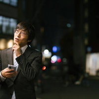 スマホで目的地を探す会社員男性、夜間の街中で迷いながら考えるの写真