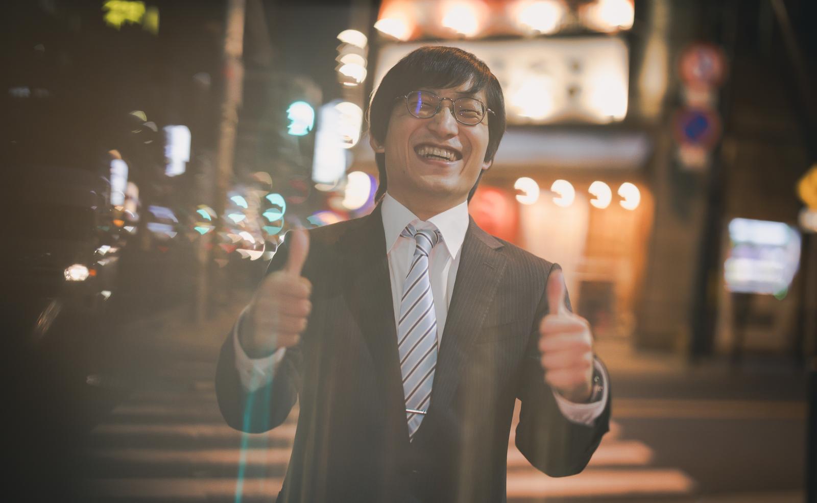 夜の街で親指を立てて笑う眼鏡をかけた会社員男性［モデル：あづま］