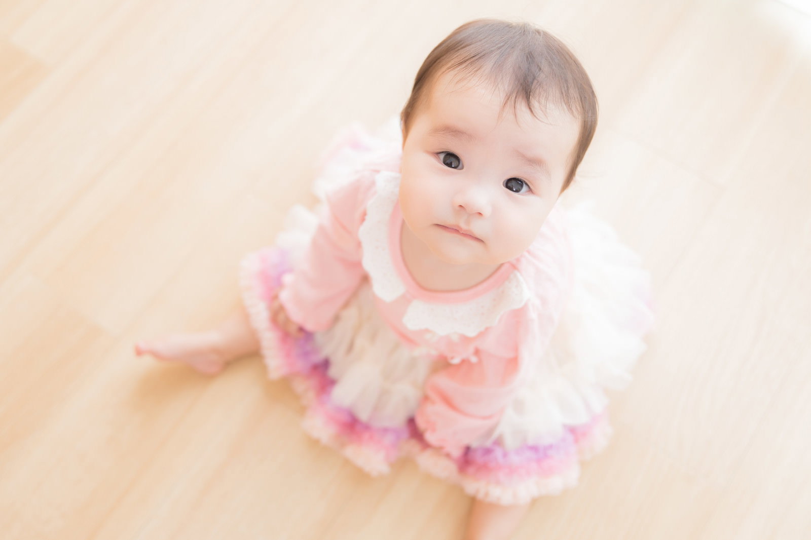 ピンク色の服を着たお座りする赤ちゃんが、上目遣いでカメラを見つめている写真［モデル：めぐな］