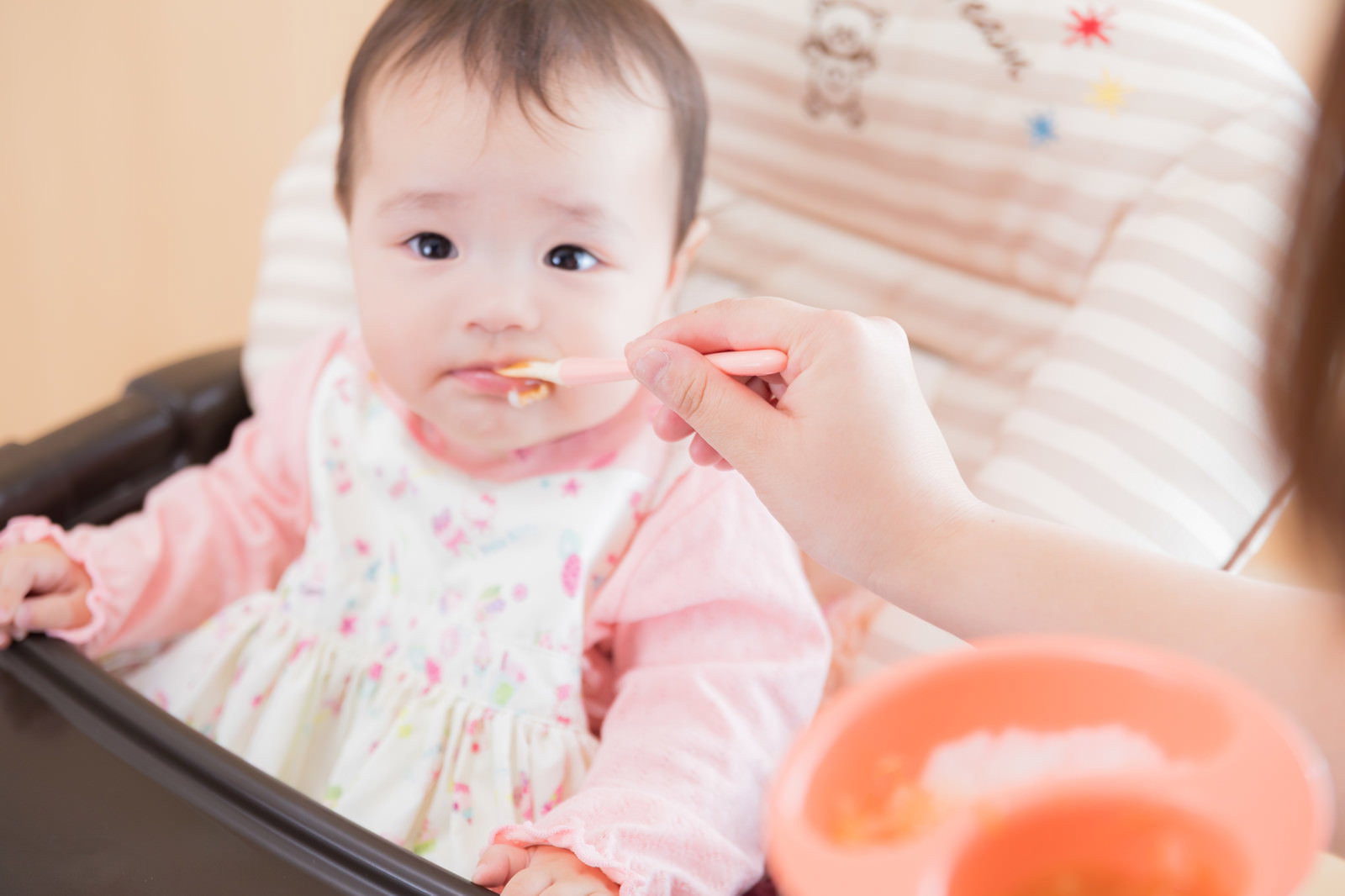 ピンクの服を着た赤ちゃんがベビーチェアで離乳食を食べている［モデル：めぐな］