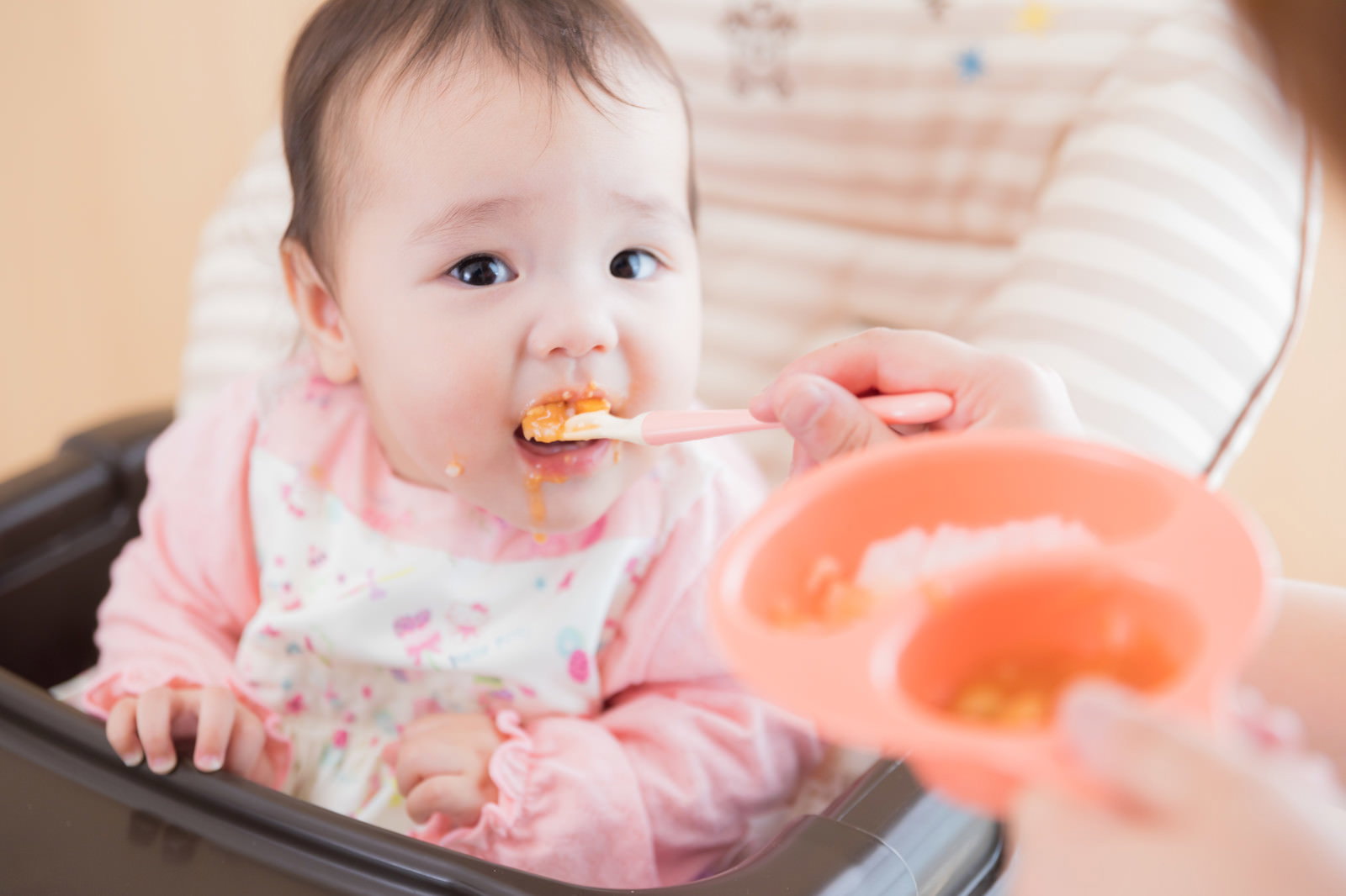 ハイチェアに座って離乳食をスプーンで食べている赤ちゃん［モデル：めぐな］