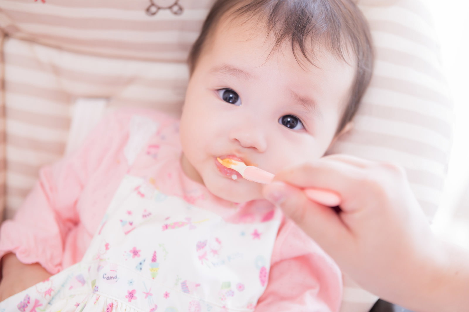 ピンクの服を着た赤ちゃんがスプーンで離乳食を食べている［モデル：めぐな］