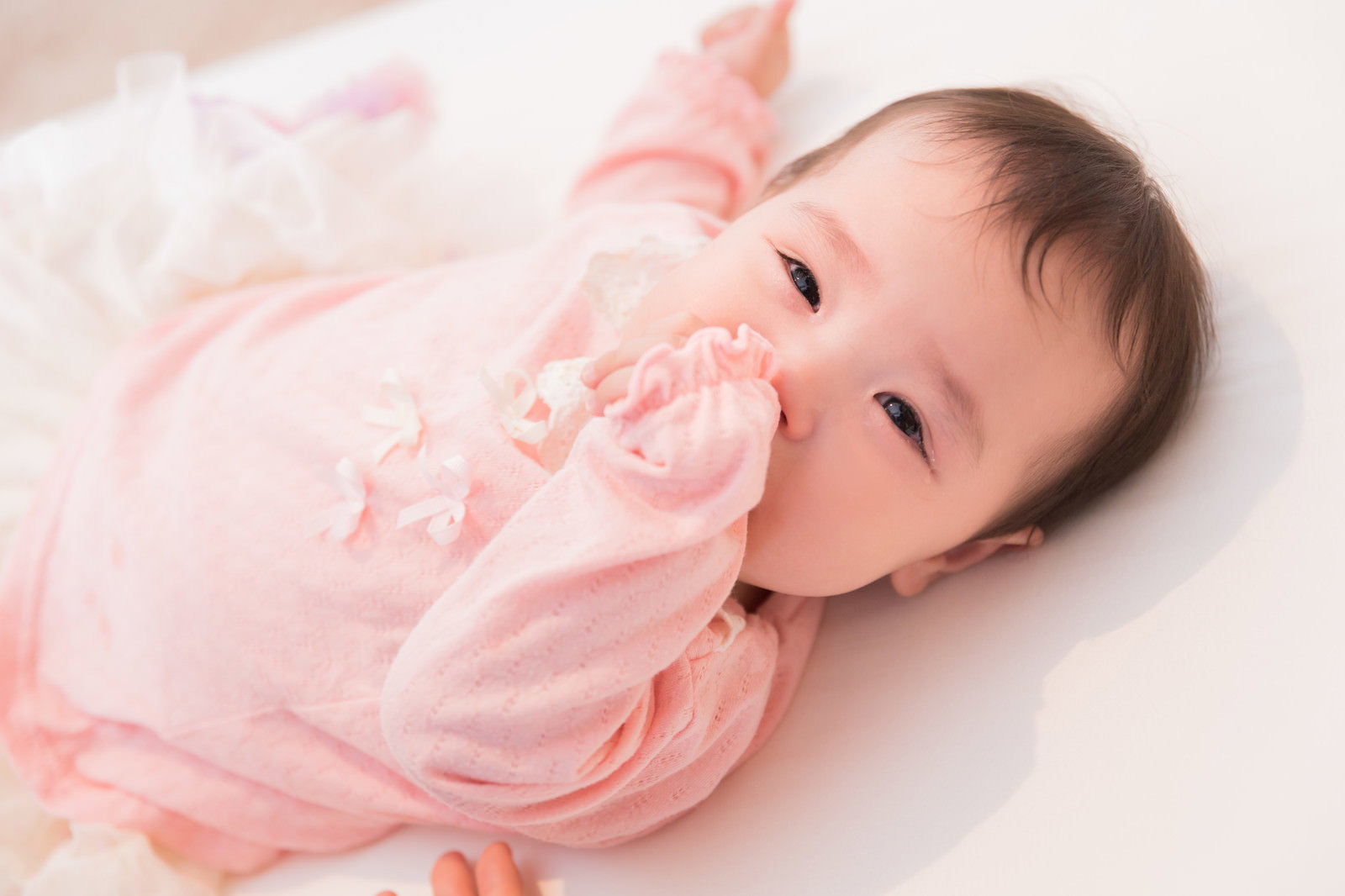 ピンクの服を着た赤ちゃんが布団の上で眠くてぐずり泣いている［モデル：めぐな］
