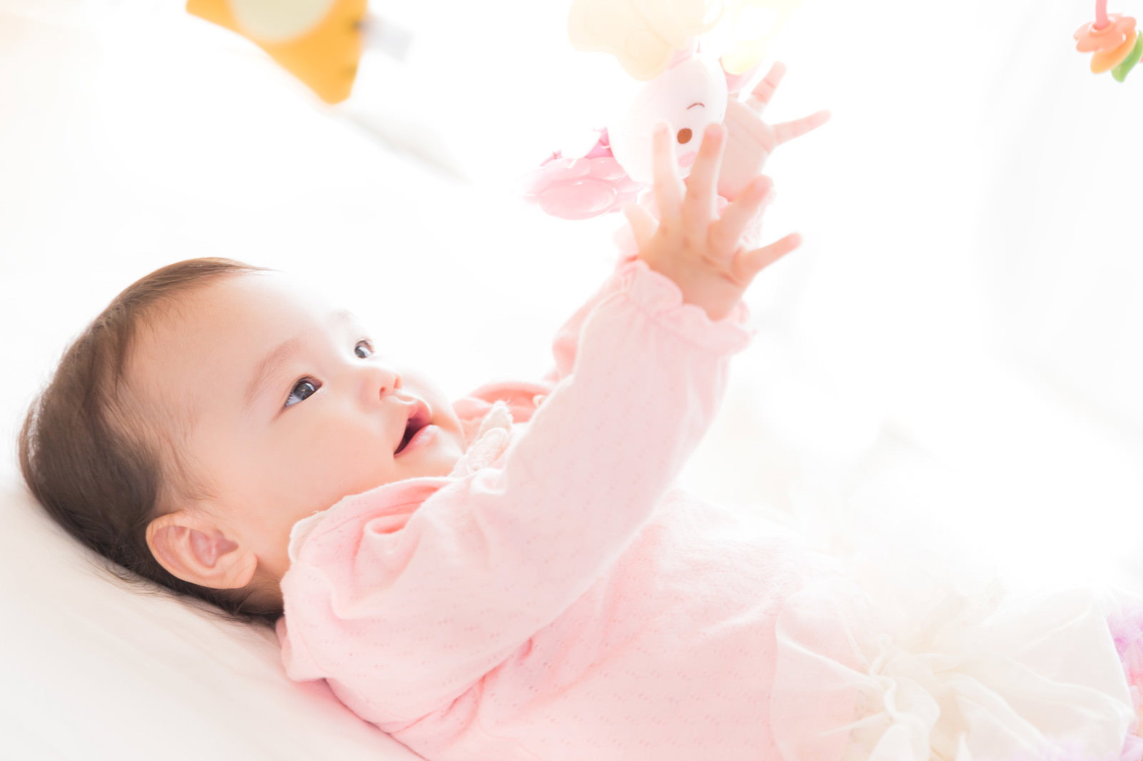 ピンクの服を着た赤ちゃんが仰向けで袖をくわえる［モデル：めぐな］