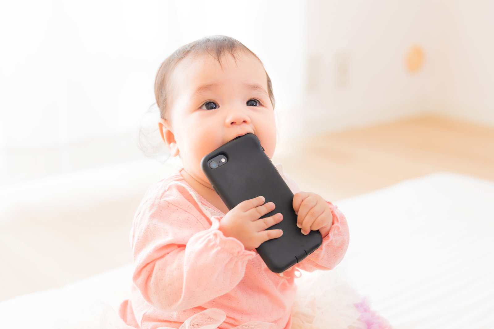 ピンクの服を着た赤ちゃんがスマートフォンを口に入れている［モデル：めぐな］
