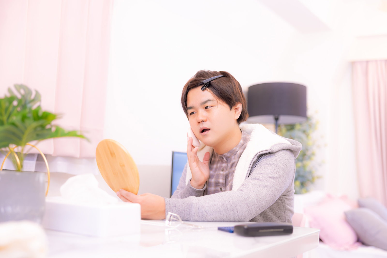部屋着を着た男性が手鏡を持ちファンデーションで化粧下地を塗っている［モデル：はしも。］