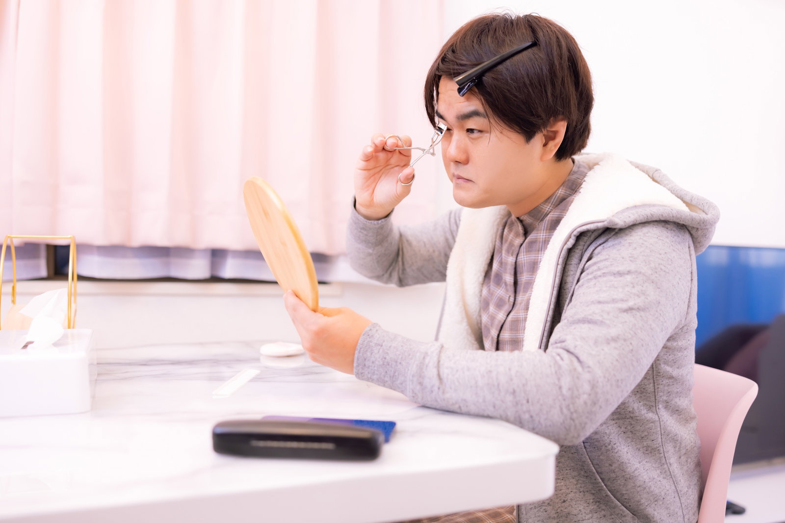 手鏡を見ながらビューラーでまつ毛をカールさせる白いパーカーの男性［モデル：はしも。］