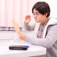 手鏡を見てビューラーでまつ毛をカールさせる男性の写真