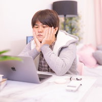 ノートパソコンの前で困惑する白いパーカー姿の男性の写真
