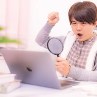 虫眼鏡を片手に持ちながらパソコンで在宅ワークする男性の写真