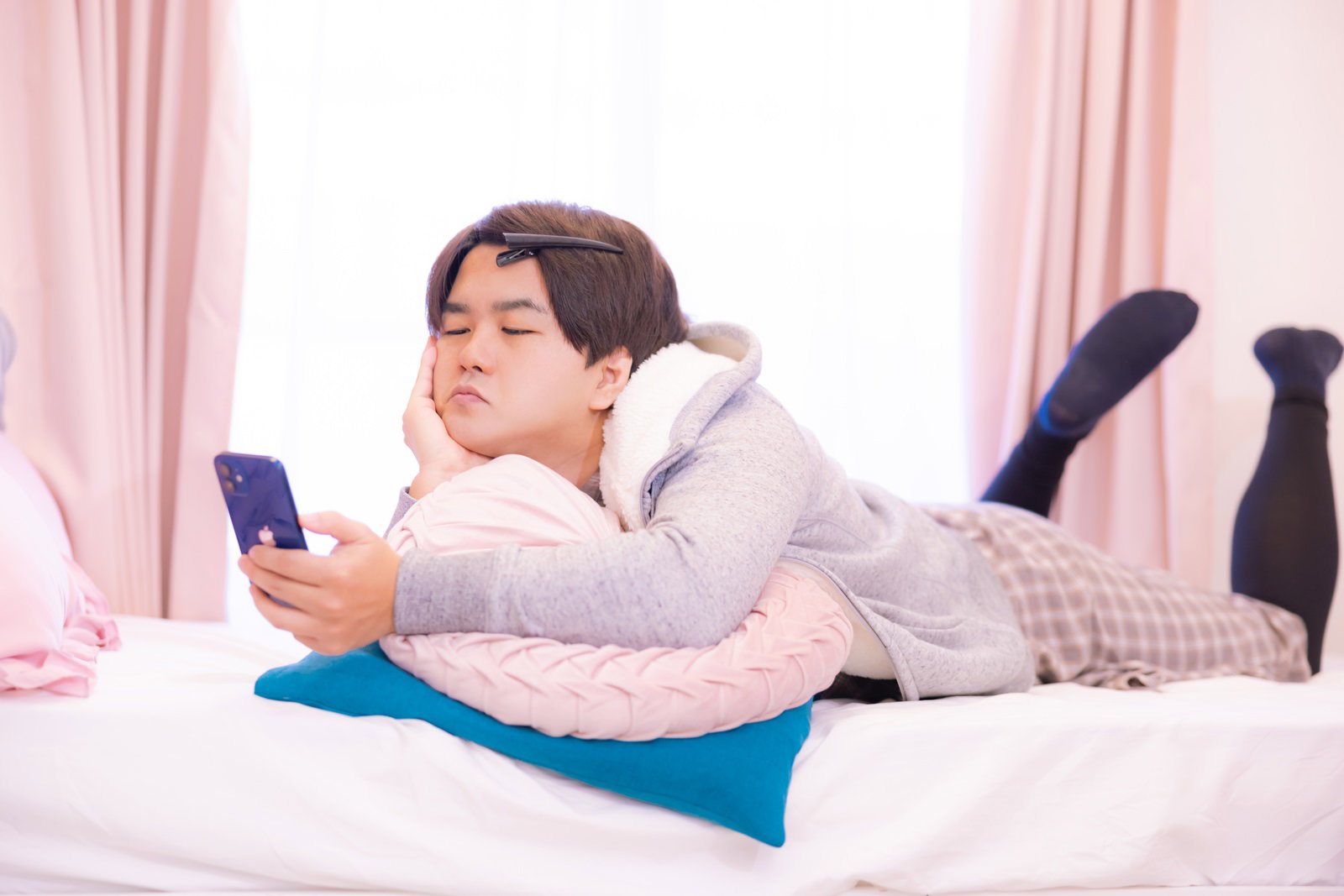 白いパーカーを着た男性がベッドでうつ伏せになりスマホを見ている［モデル：はしも。］