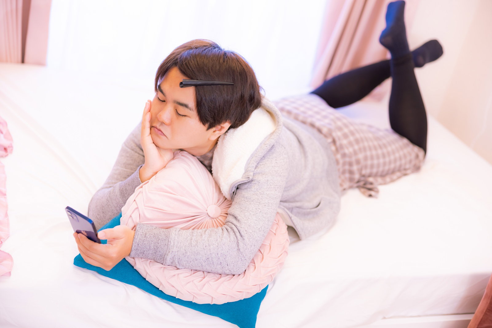 白いシャツを着た男性がベッドに横たわりスマホを見ている［モデル：はしも。］