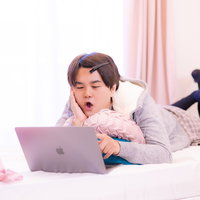 部屋着でベッドに横たわりネットサーフィンする男性の写真