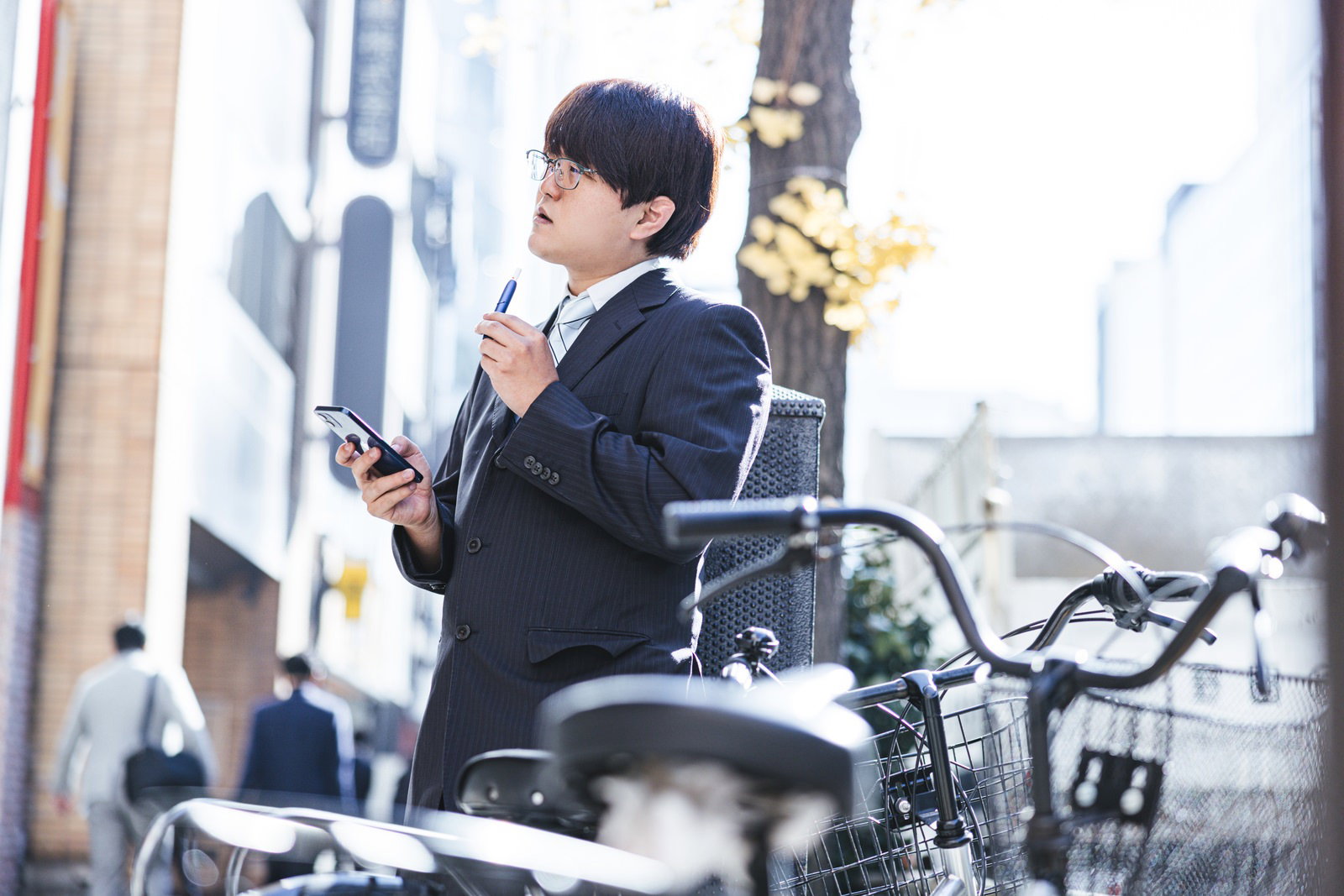 黒いスーツを着た会社員が電子タバコを持ちながらスマートフォンを確認している［モデル：はしも。］
