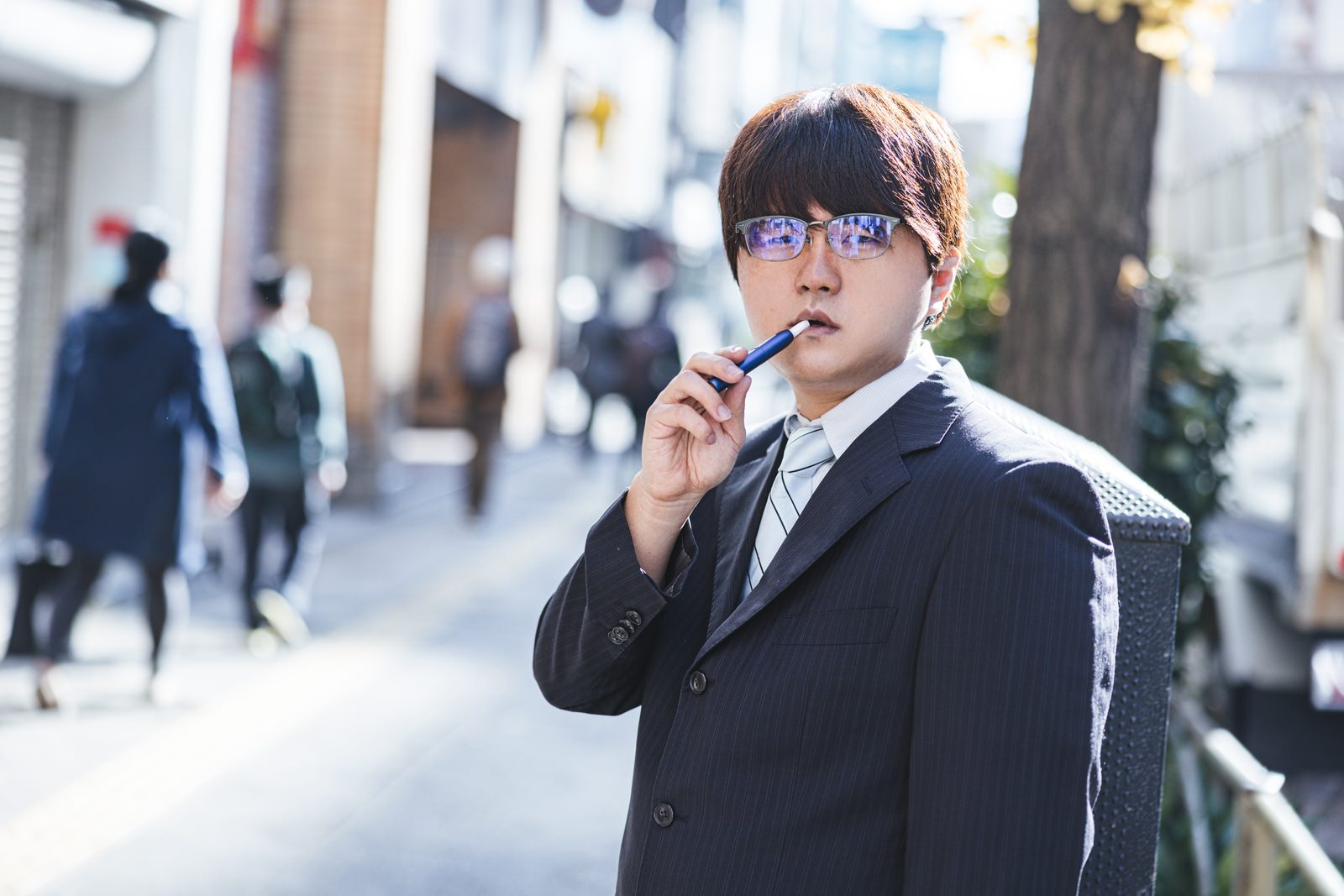 路地で電子タバコを持ち不機嫌な表情を見せるスーツ姿の会社員男性［モデル：はしも。］
