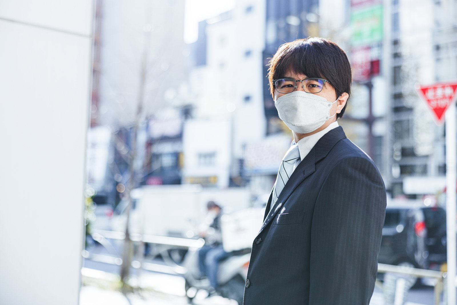 街中で振り返るマスクを着用した黒いスーツ姿の男性会社員［モデル：はしも。］