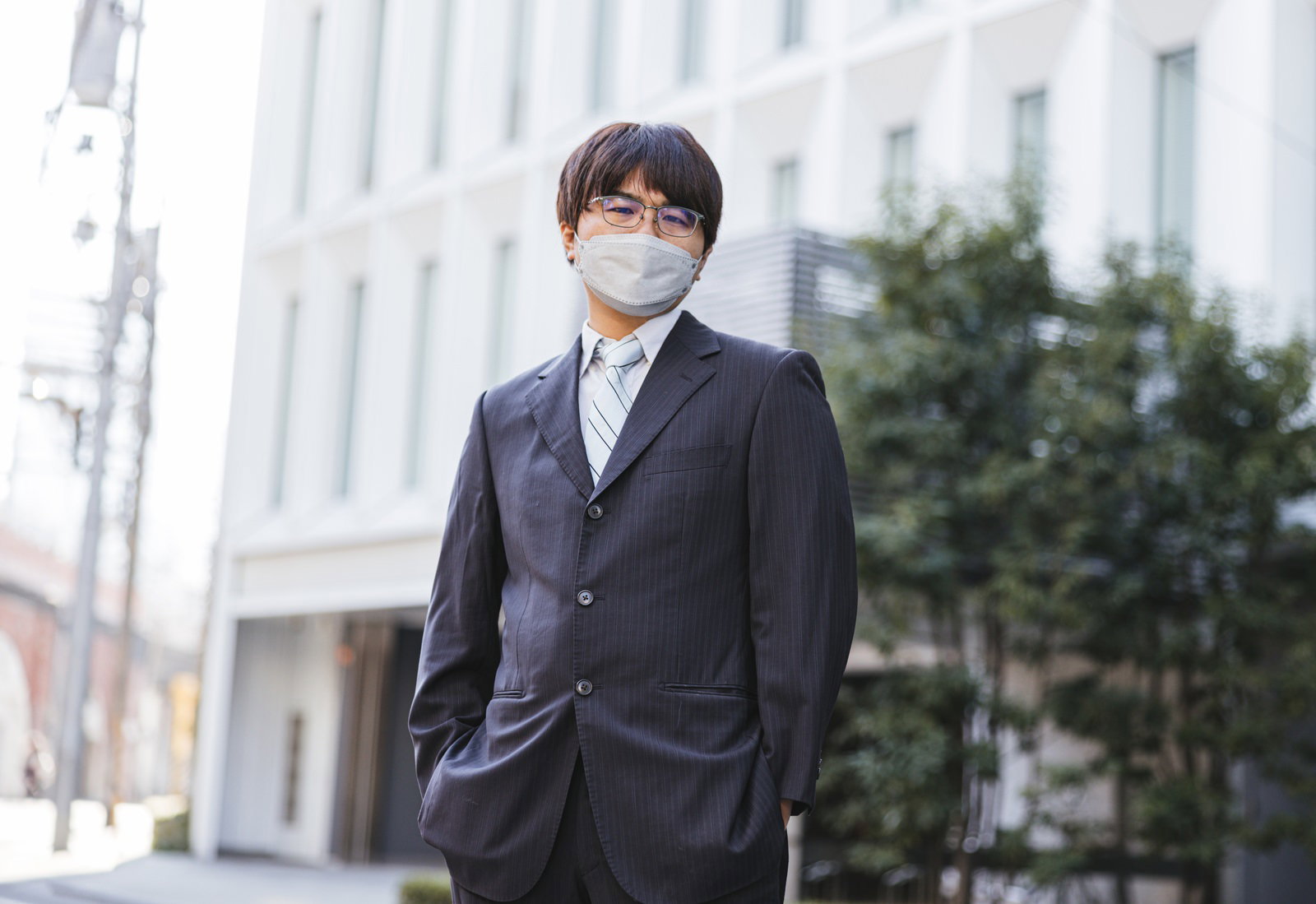 マスクをしたスーツ姿の若い会社員がビル前に立つ［モデル：はしも。］