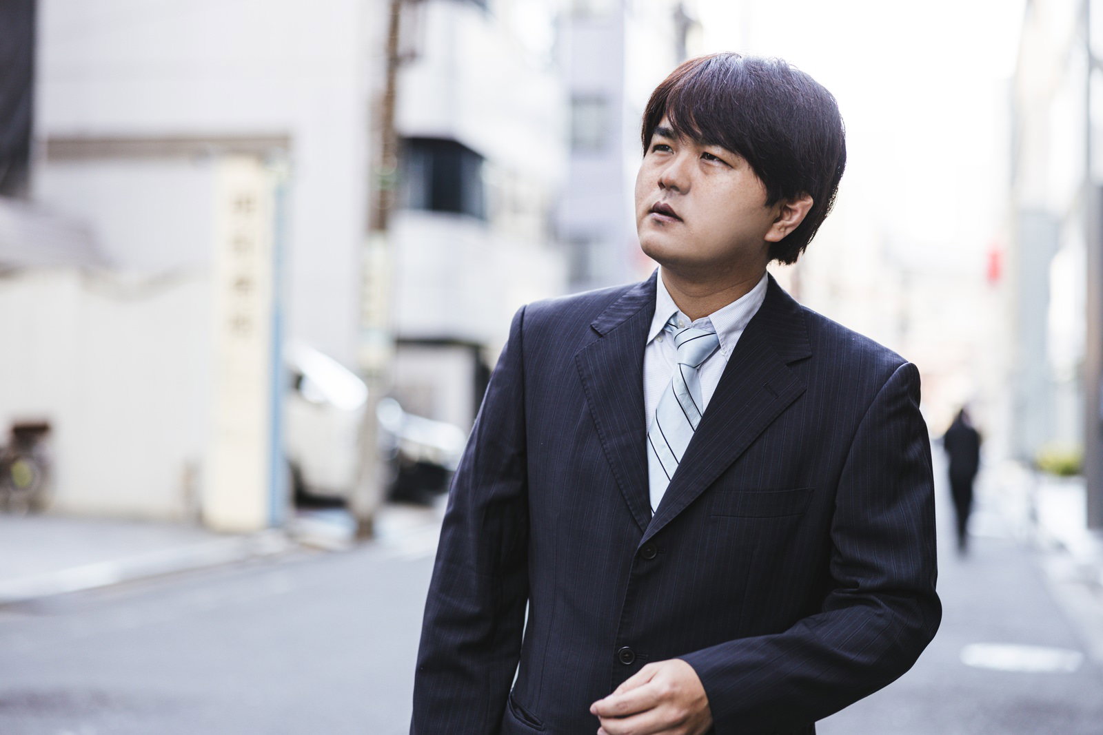 黒いスーツを着た男性が街角で張り込みをしている様子［モデル：はしも。］