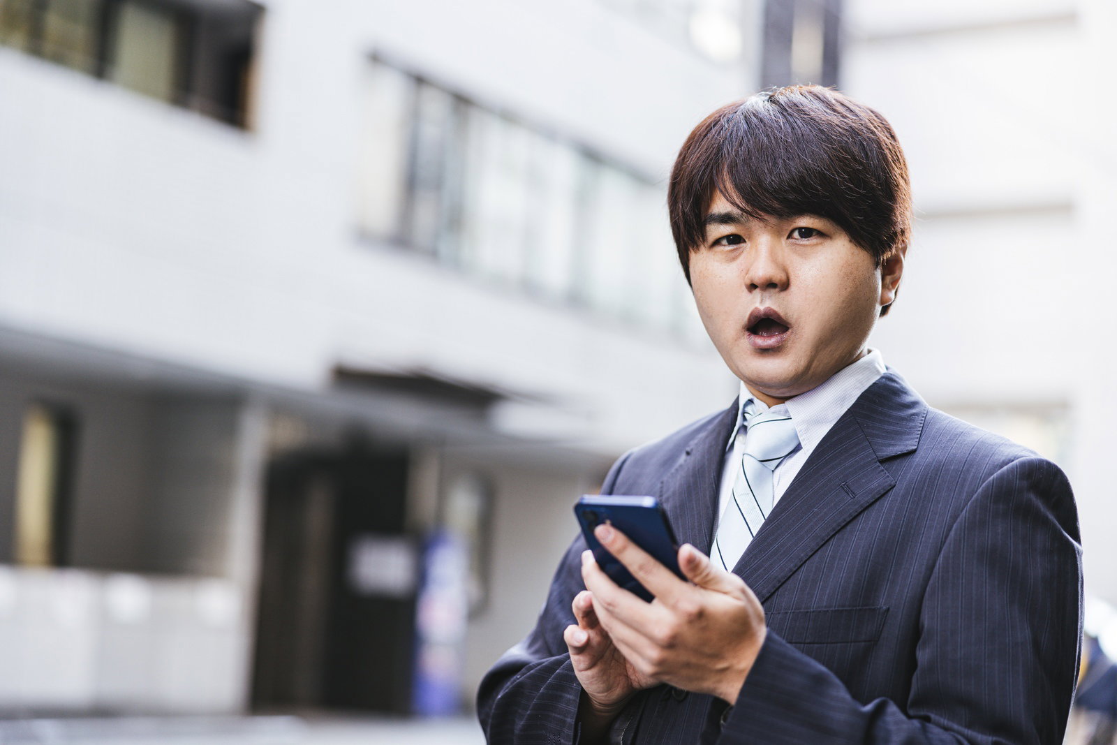 屋外でスマートフォンを見て驚いた表情を浮かべるスーツ姿の会社員男性［モデル：はしも。］