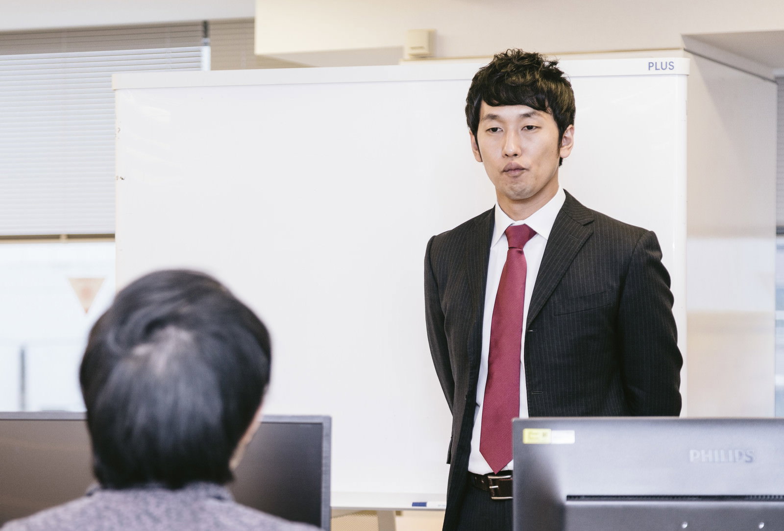 会議室のホワイトボード前に立つスーツ姿の男性。赤いネクタイを着用し真摯な表情で立っている［モデル：大川竜弥］