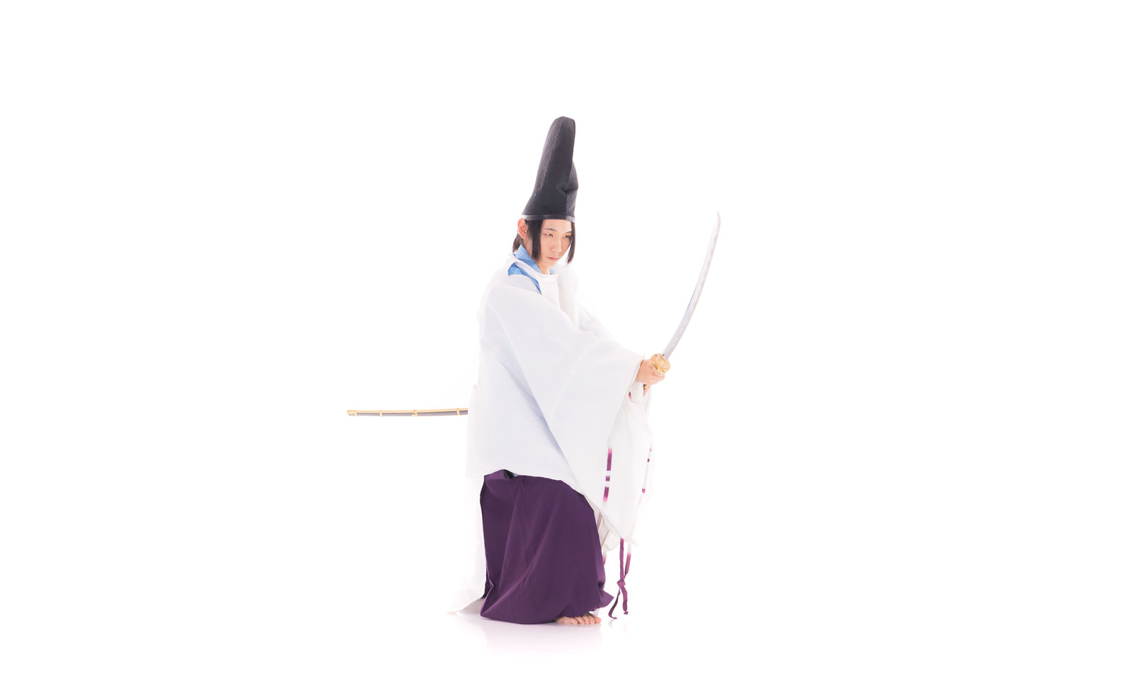 白い狩衣と黒い帽子を身につけた男性が、刀を握って腰を落とした構えを取っている［モデル：まーこ］