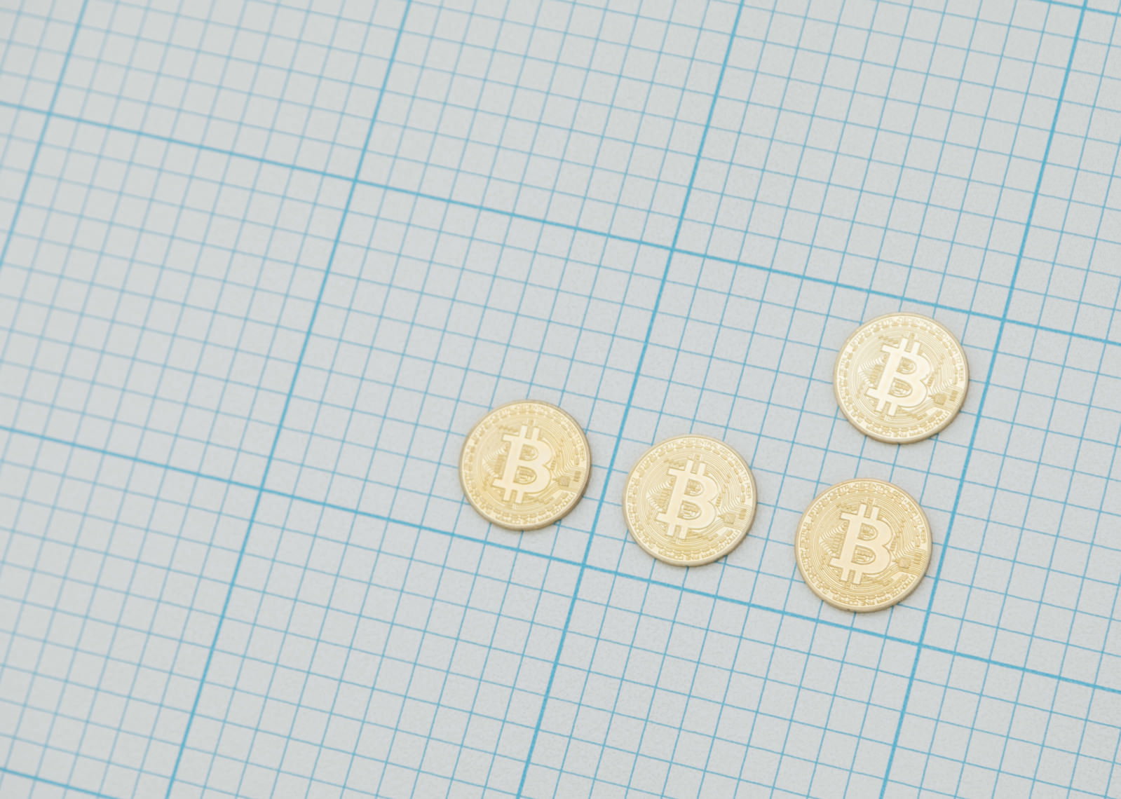 青い方眼紙の上に金色のビットコイン硬貨が4枚置かれている