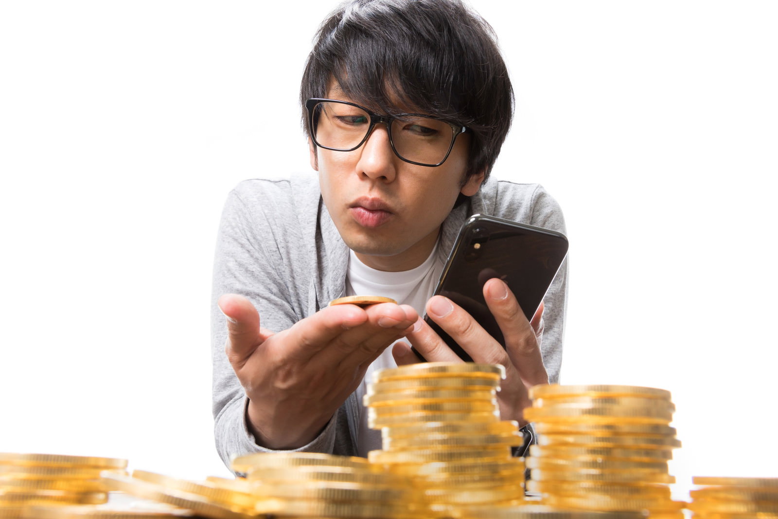 金色のコインが積み重ねられた前でスマートフォンを見つめる眼鏡の男性投資家［モデル：大川竜弥］