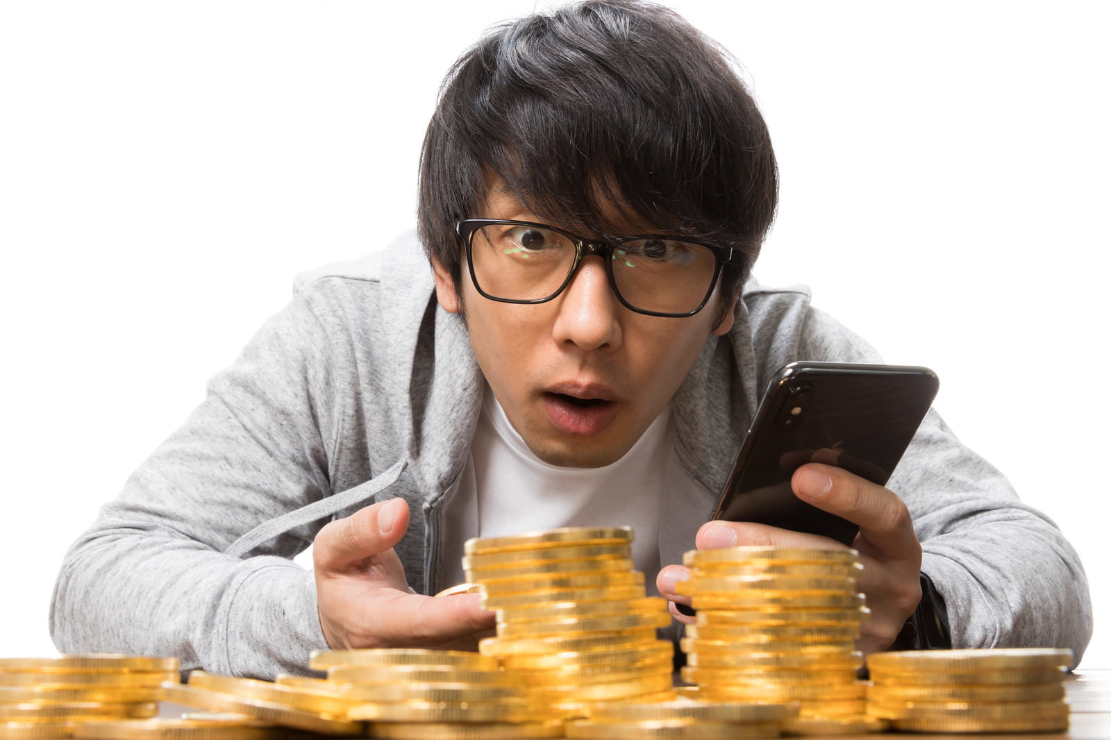 スマホを握る男性と積み重ねた金貨。前かがみで唖然とした表情。［モデル：大川竜弥］