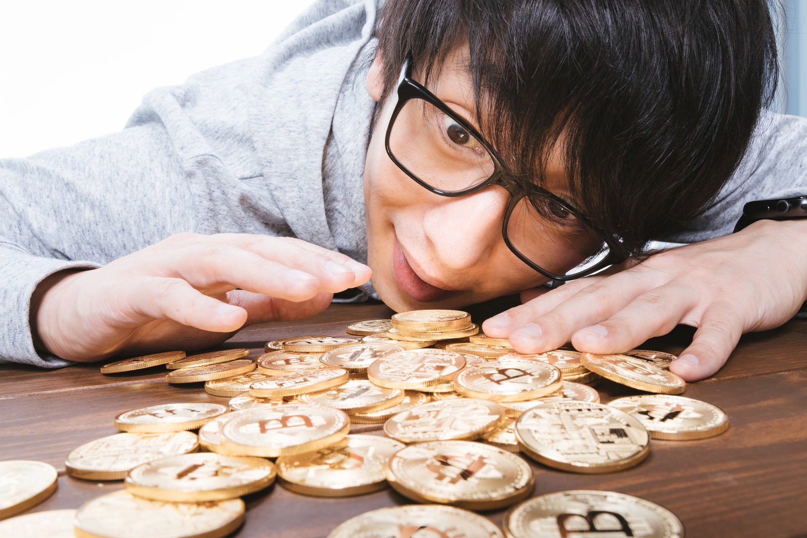 眼鏡をかけた男性が机の上の金色のビットコインコインを身を乗り出して見つめている［モデル：大川竜弥］