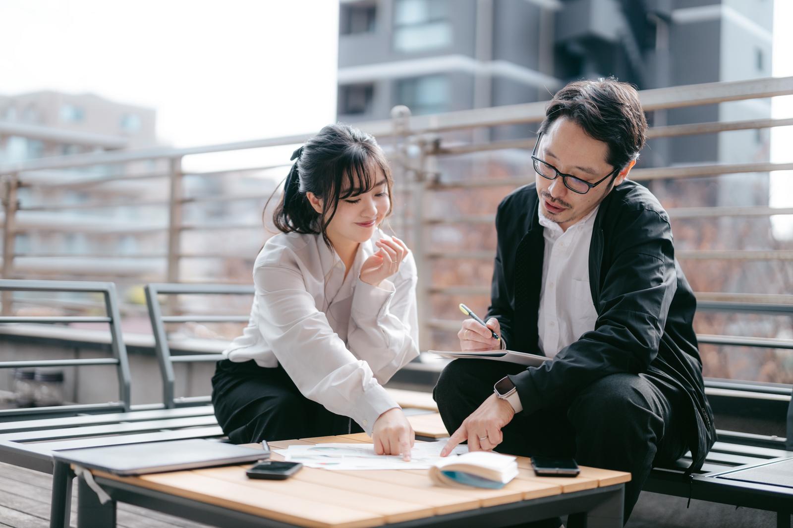 屋上テラスで資料を確認する眼鏡をかけた男性と白シャツの女性［モデル：河村友歌 ゆうせい］