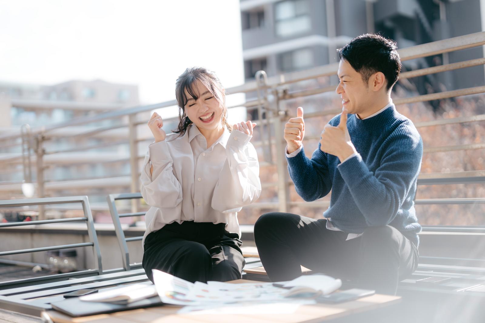屋上で打ち合わせしながら喜ぶ男性と女性社員［モデル：モデルリリース 河村友歌］