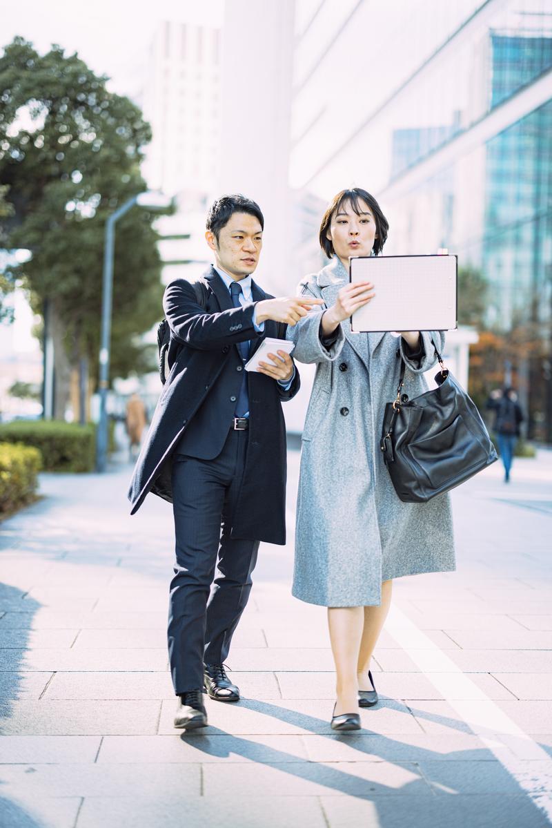 都市の歩道でタブレットを持って打ち合わせする男女のビジネスパーソン［モデル：たけべともこ モデルリリース」］