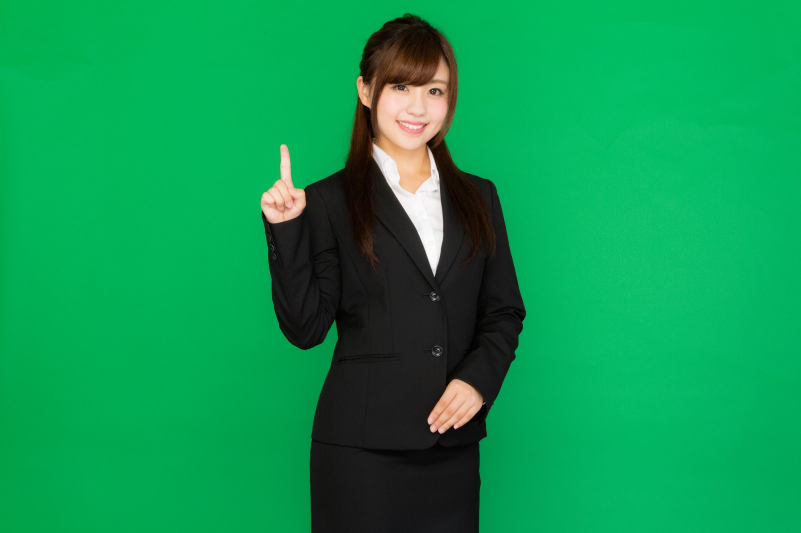 グリーンバックの前で黒スーツを着た女性が人差し指を立てて上を指し、笑顔で正面を向いている［モデル：河村友歌］