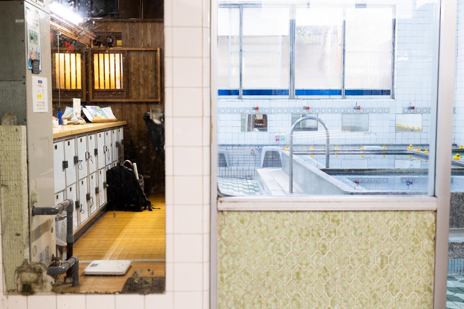 A retro sento bath area visible through a glass door, with lockers and a weighing scale lined up in the changing room in the foreground
