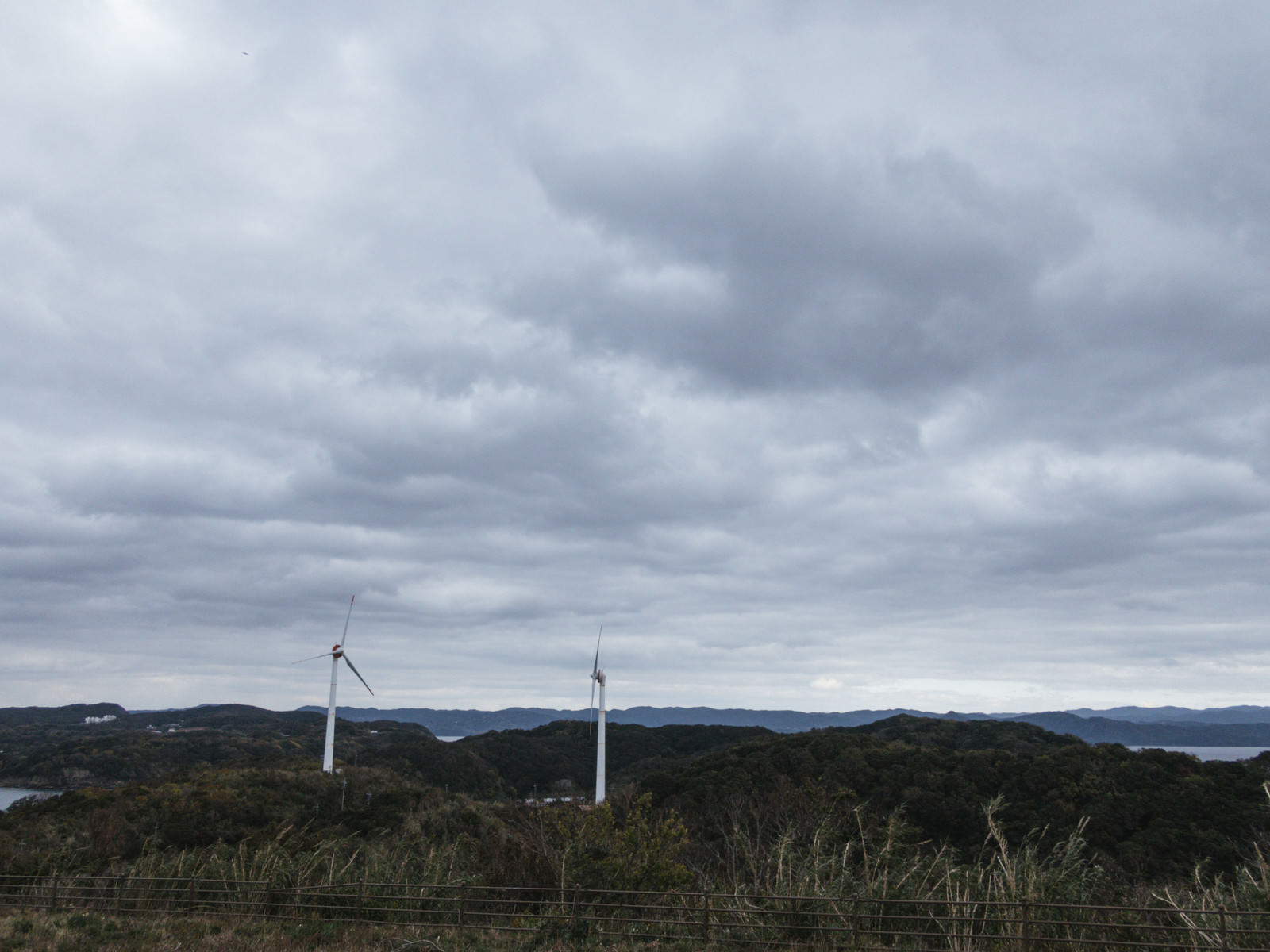 曇り空の下、山中に設置された風力発電機を展望台から眺めた景色