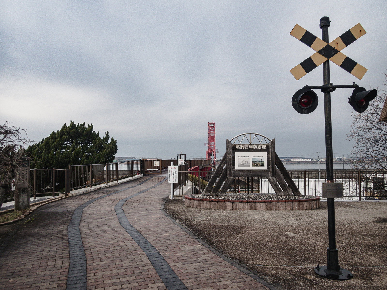 曇り空の下、廃駅跡の遊歩道に立つ踏切信号と記念碑、背景に赤い橋塔が見える