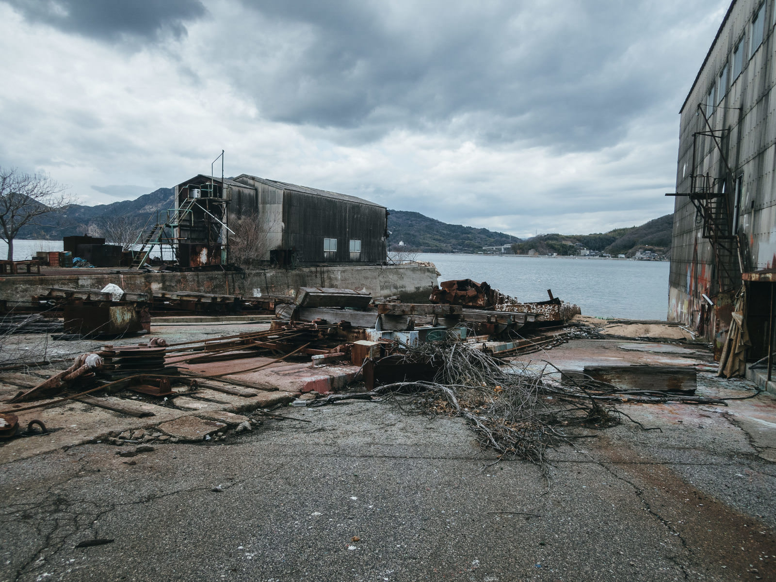 曇り空の下、海沿いに建つ造船所の工場建物と倉庫、散乱した木材