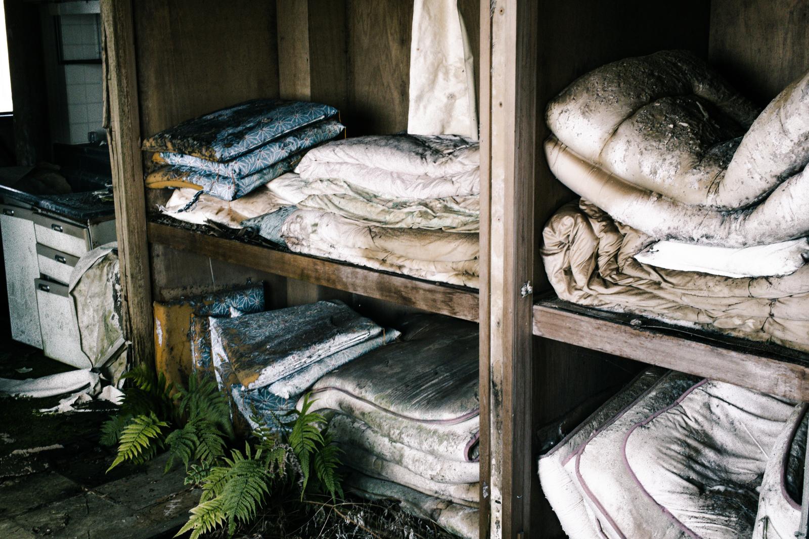 薄暗い廃墟の店舗カウンター。ガラス戸棚に食器が並び、天板に古い瓶などが散乱している。