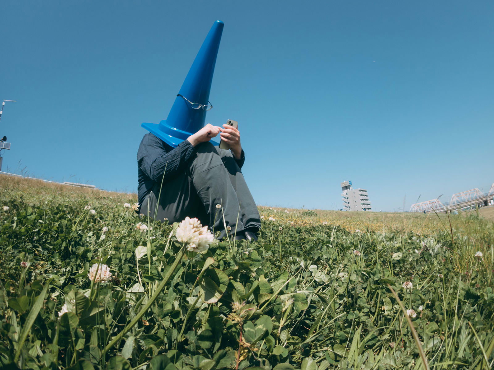 河川敷の草原でオレンジ色の三角コーンを被った人物がスマートフォンを操作している