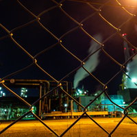 金網越しに見るオレンジ色の照明が照らす工場夜景の写真