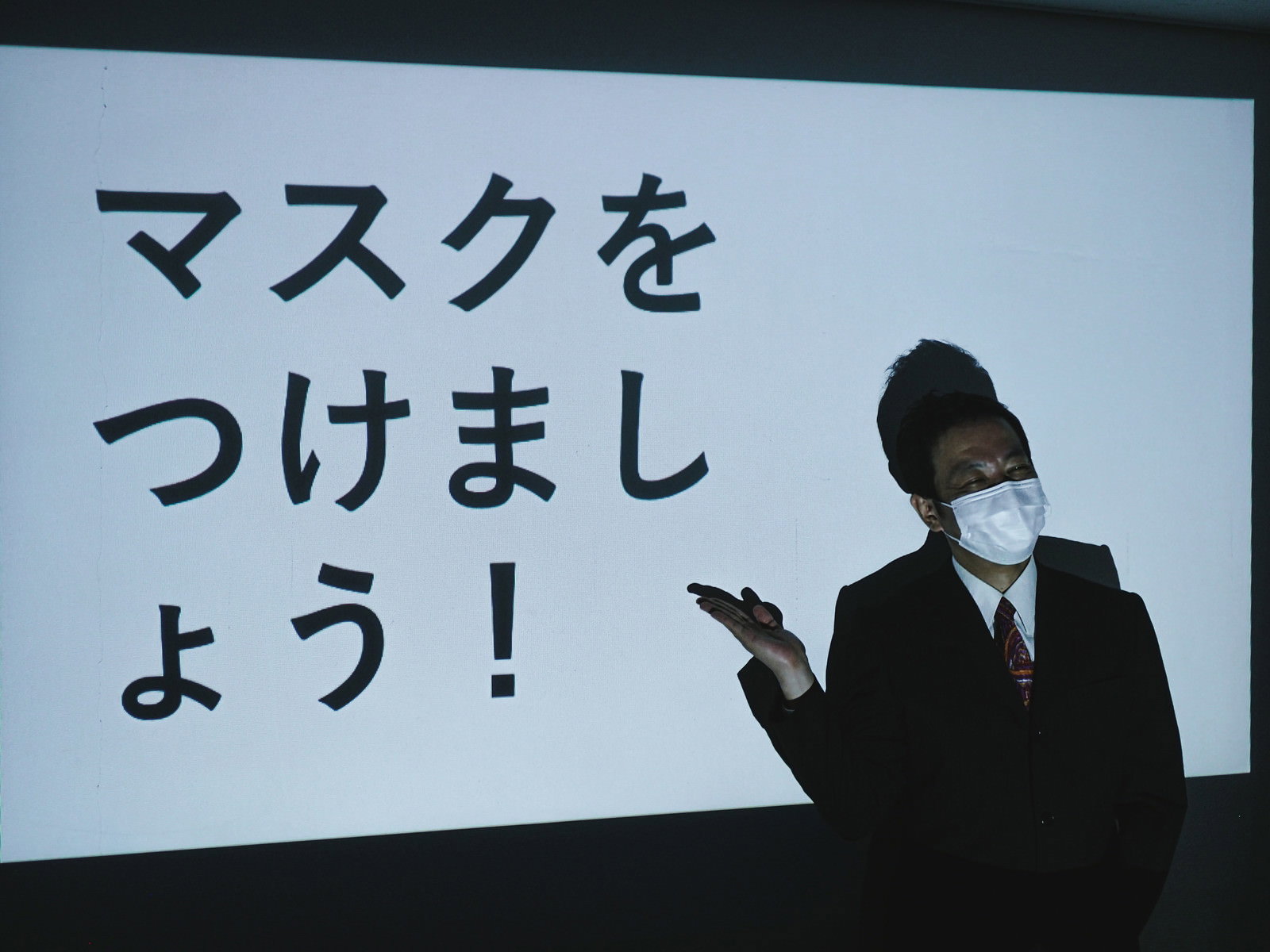 マスク着用を呼びかけるスライドの前で説明する男性［モデル：のせさん］