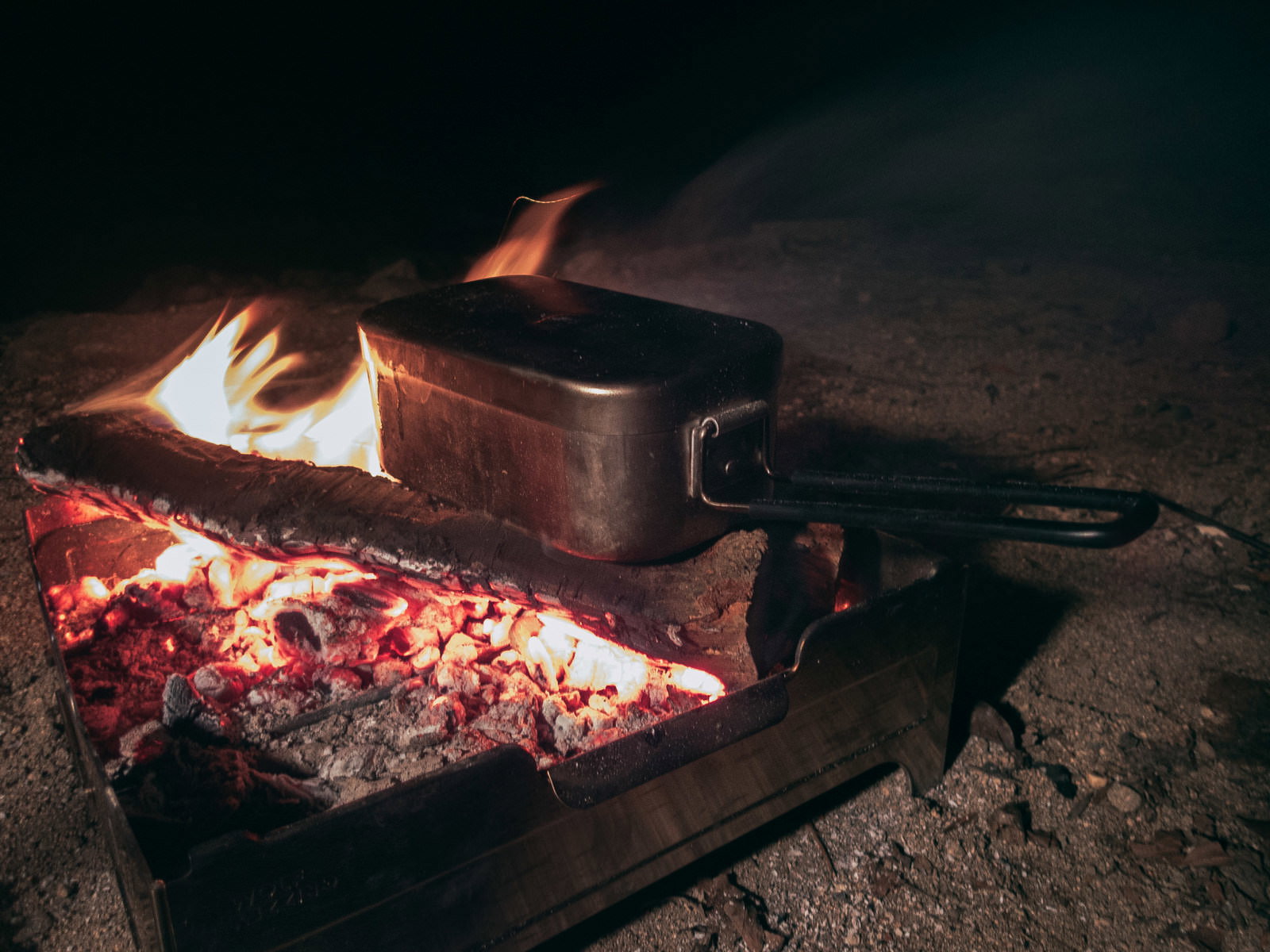 焚き火の上で炊飯中の飯盒、赤い炎に照らされる夜のキャンプシーン