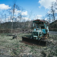 立ち枯れた木々が残る山道に放置された青いブルトーザー廃車の写真