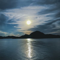 曇り空に浮かぶ太陽とレイラインが水面に映る風景の写真